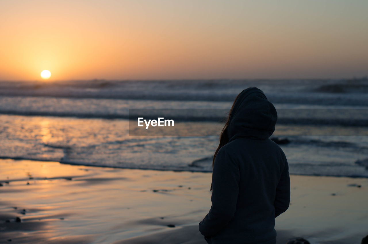 Rear view of woman standing at beach during sunset