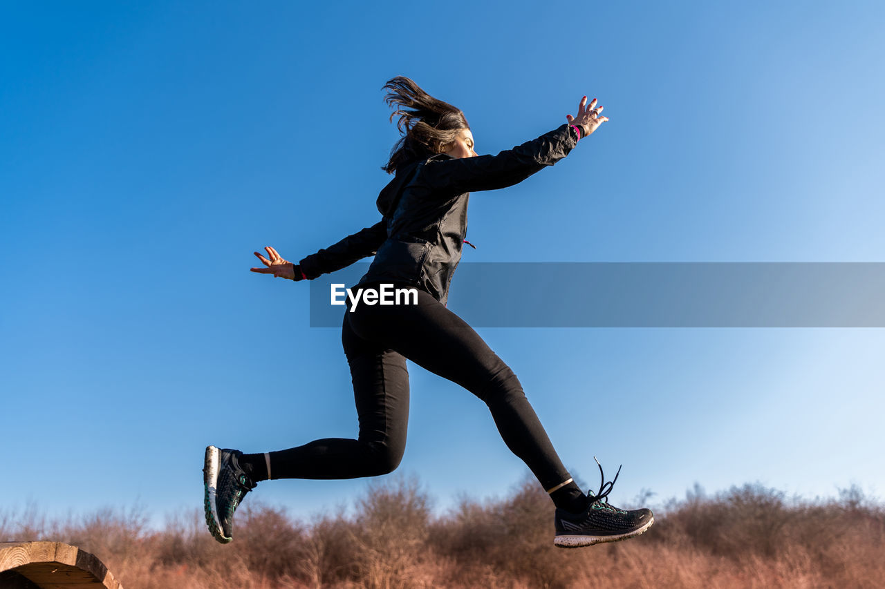 Young female runner is jumping over wooden hurdles in countryside - concept of health and wellness