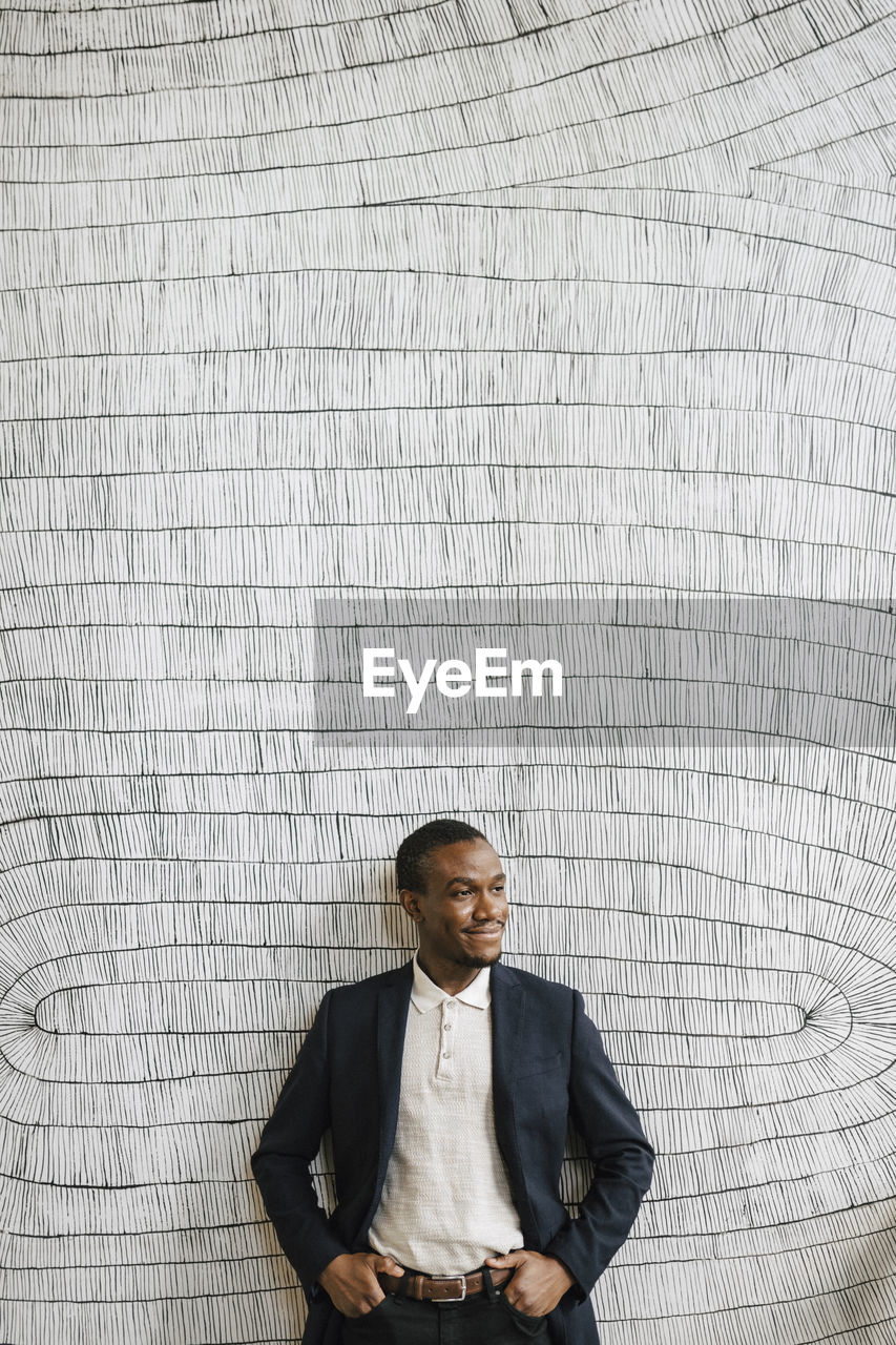 Smiling male professional standing with hands in pockets in front of wall