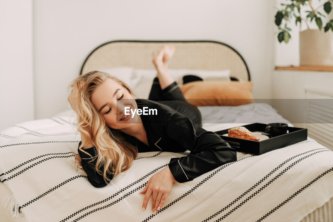 A happy pretty young woman in pajamas is resting and having breakfast in bed in a cozy apartment