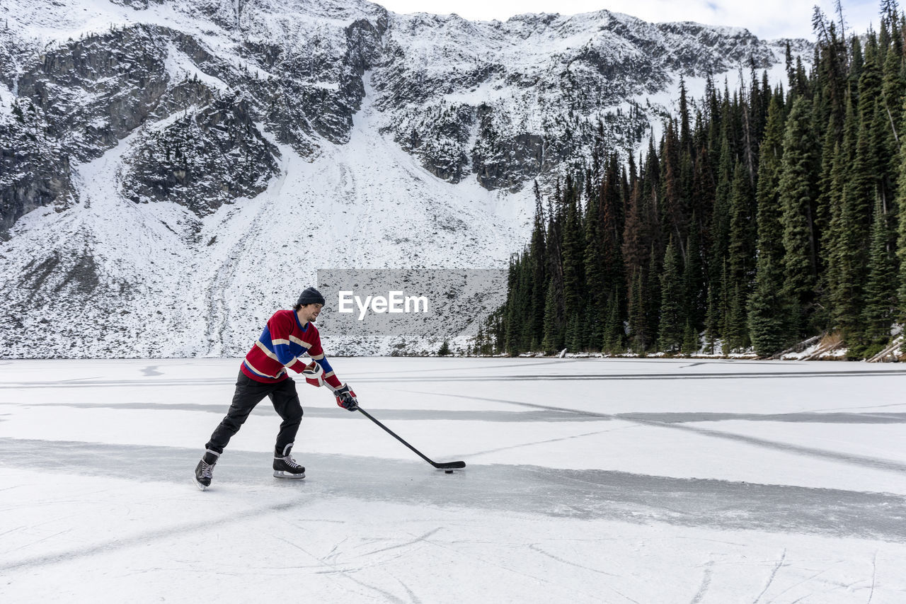 Young man playing ice hockey on lake near snow covered mountain