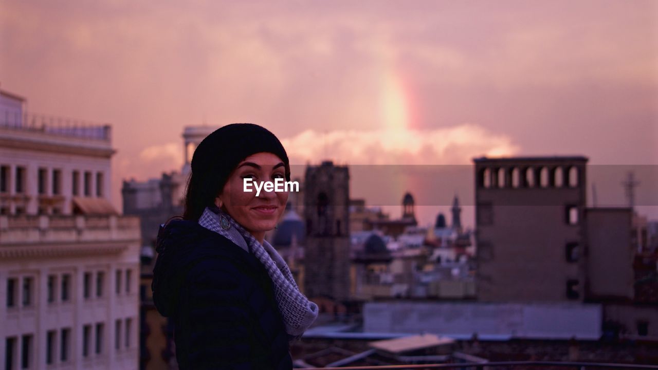 Portrait of young woman standing against cityscape during sunset