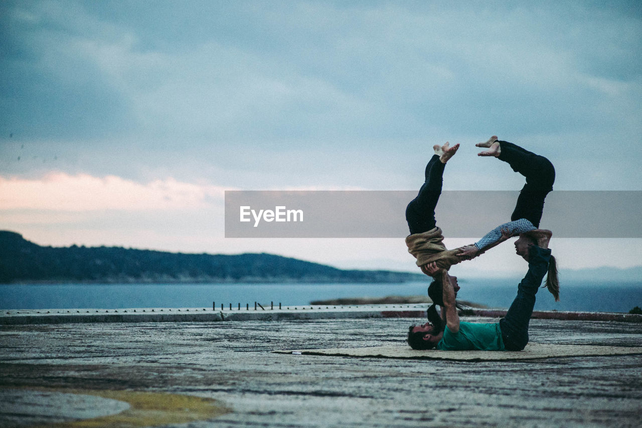 Full length of man balancing women on feet by sea against cloudy sky