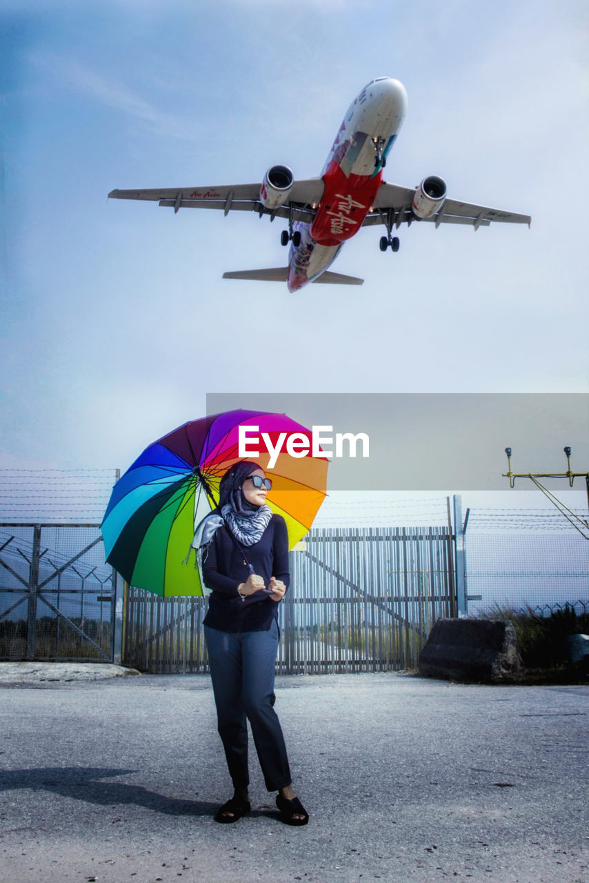 Full length of woman standing while holding umbrella against airplane in sky