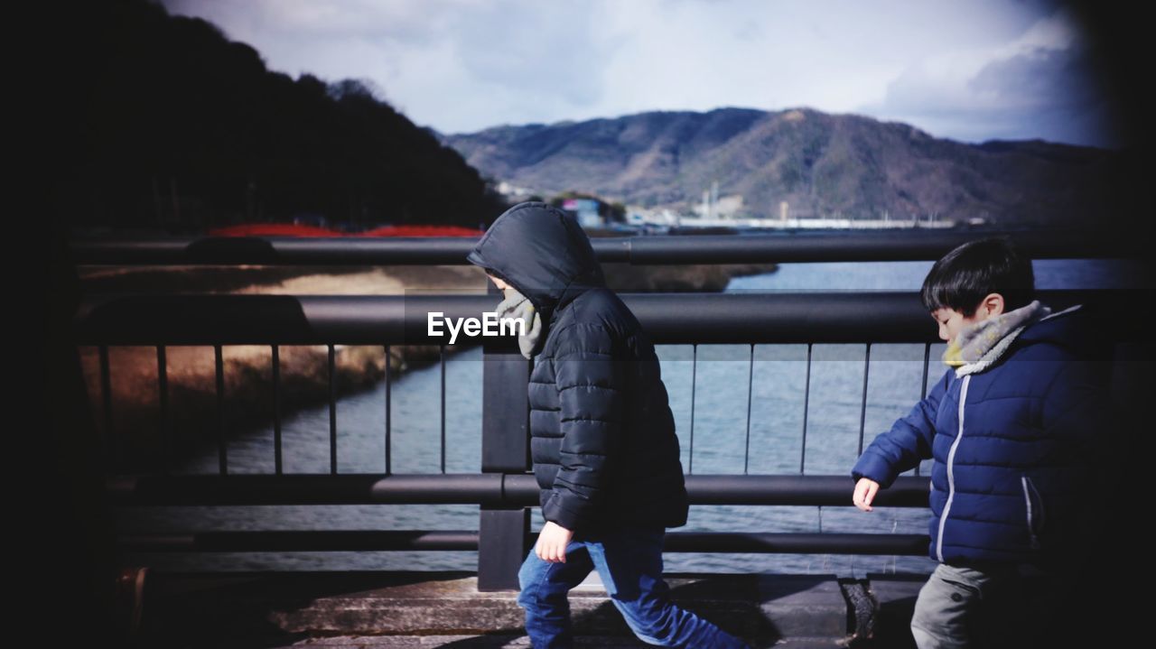 Side view of boys wearing warm clothing walking on bridge over river