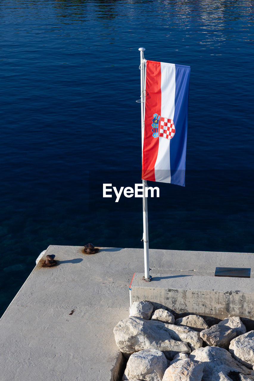 flag, water, patriotism, nature, sea, no people, day, blue, rock, outdoors, sunlight, tranquility, high angle view, red, symbolism, pole, independence, rippled