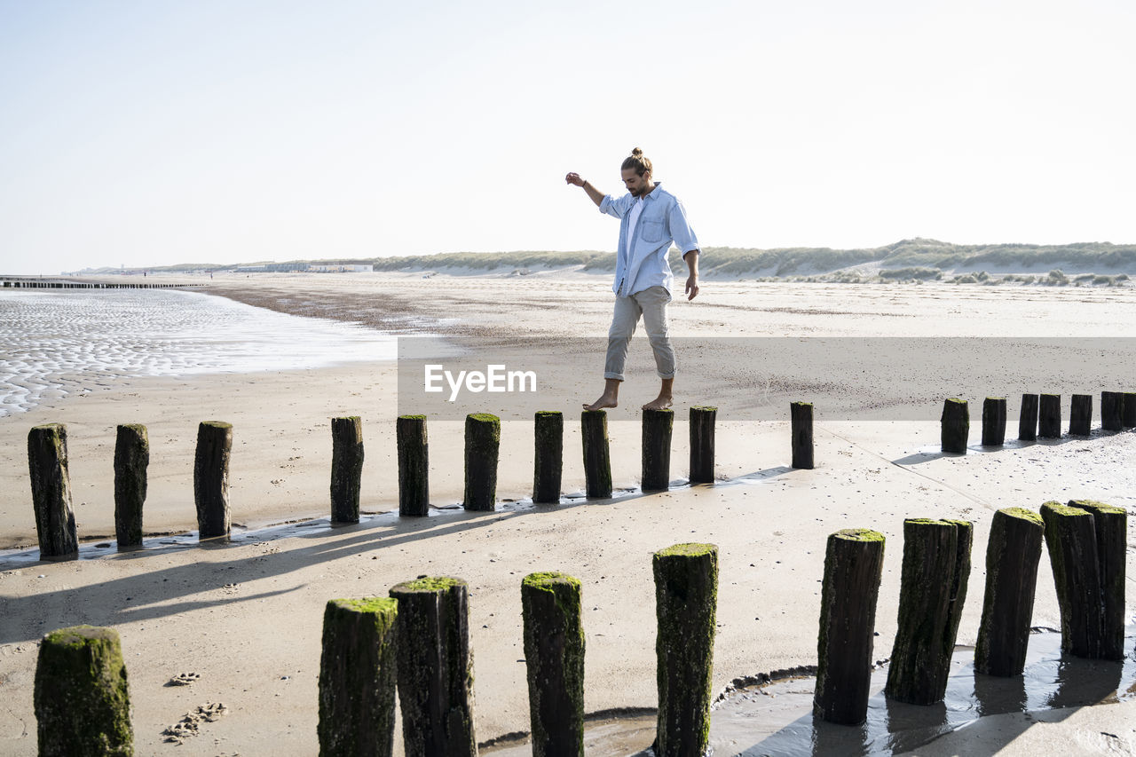 Young man walking on wooden posts at beach against clear sky