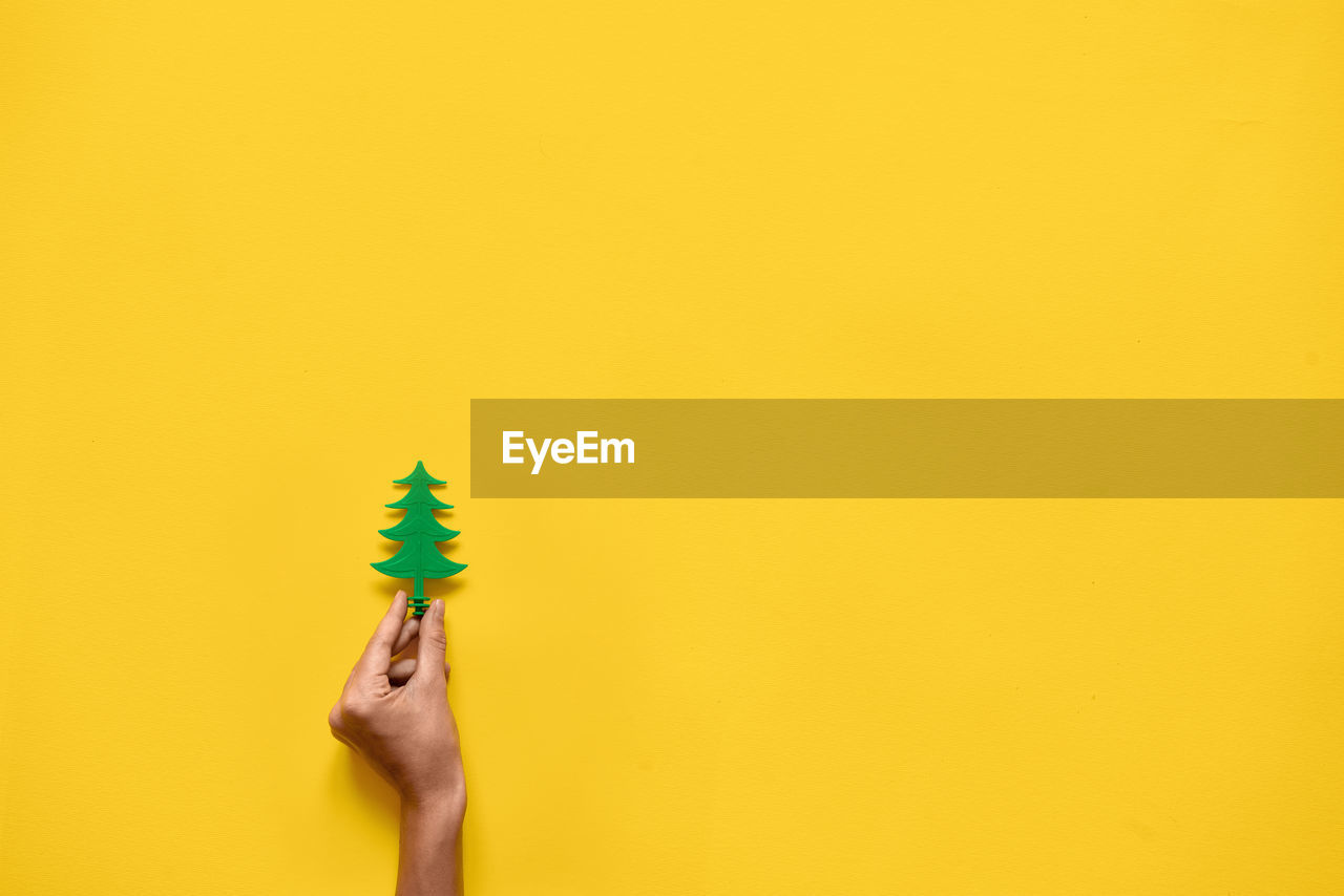 Cropped hand of person holding small plastic tree against yellow background