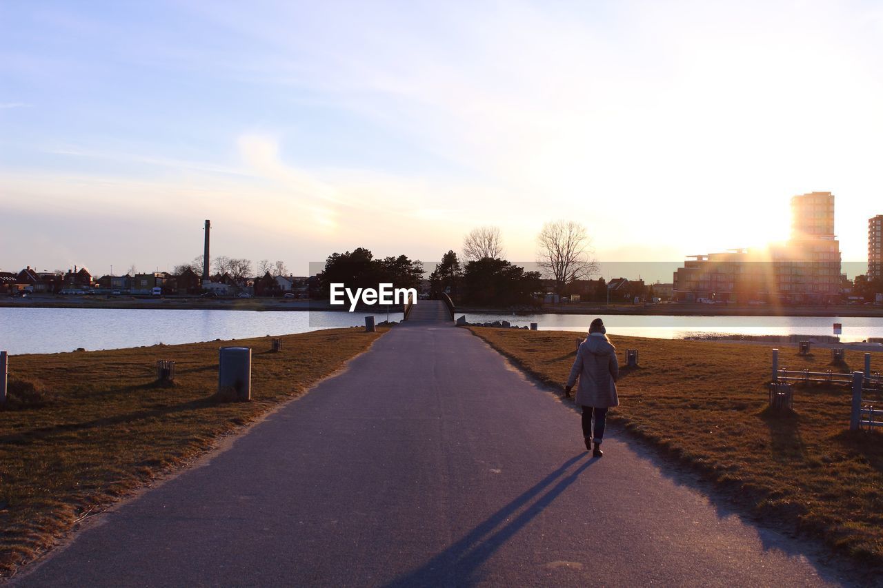 REAR VIEW OF WOMAN WALKING ON ROAD IN CITY AGAINST SKY