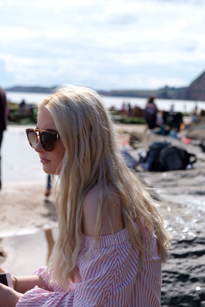 Young woman wearing sunglasses sitting at shore of beach against sky