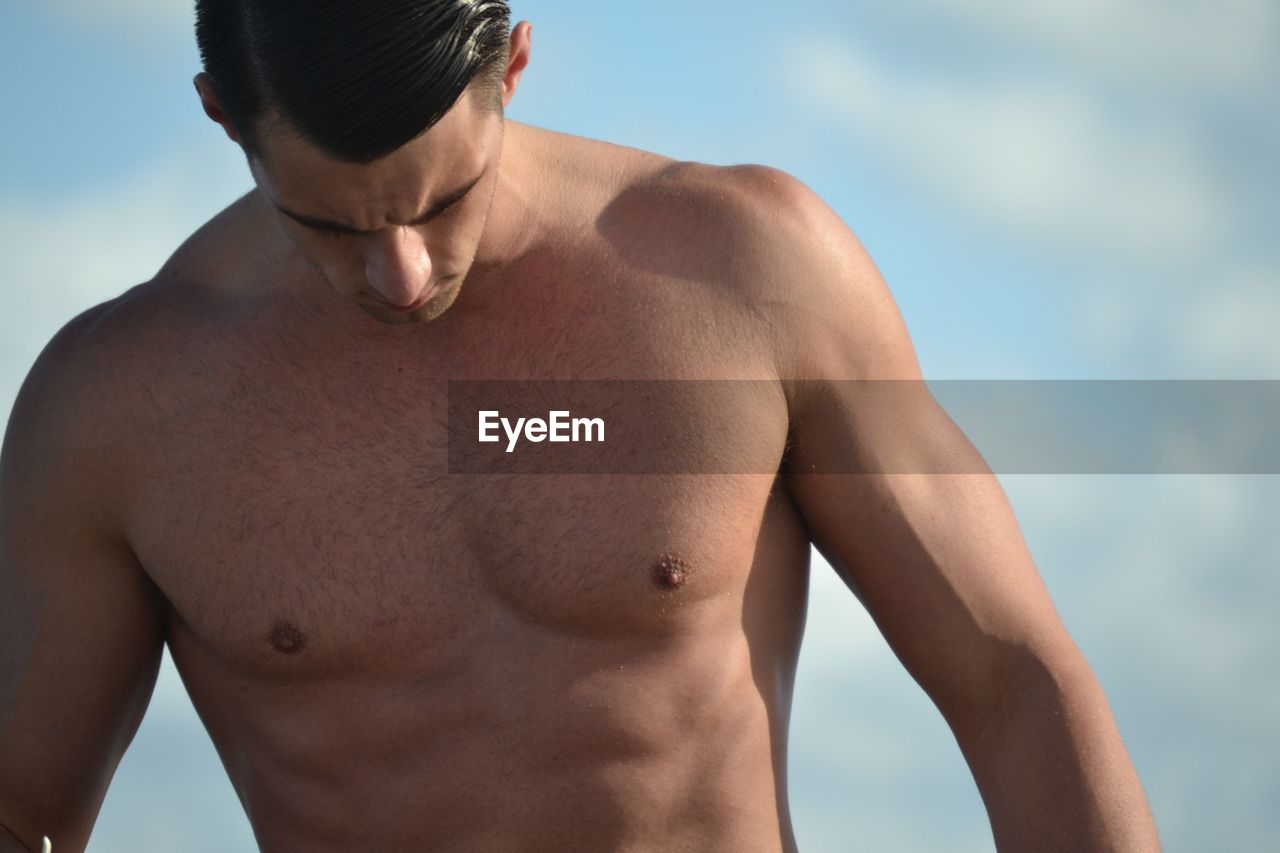 Shirtless man looking down while standing against sky