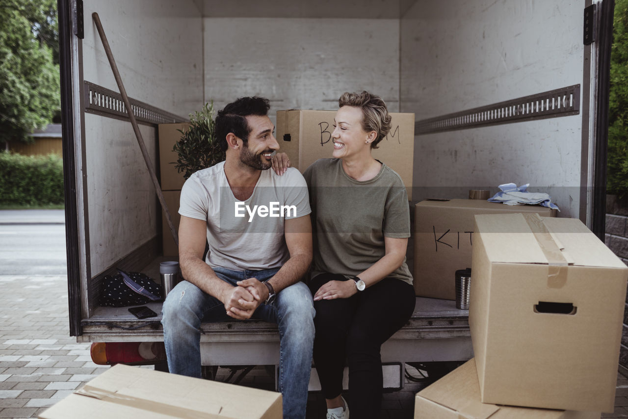 Smiling male and female partners looking at each other while sitting by cardboard boxes in van