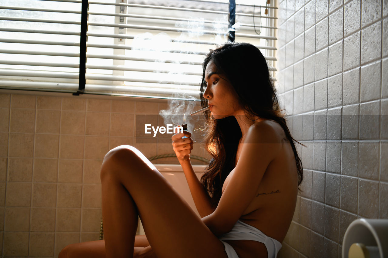 Side view of shirtless young woman lighting cigarette in bathroom