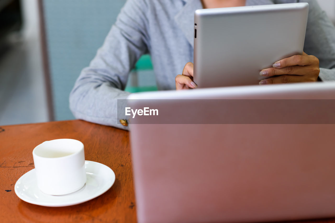 Close-up of coffee cup on table while woman using digital tablet