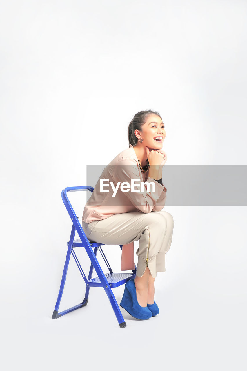 Smiling fashionable woman sitting on ladder against white background