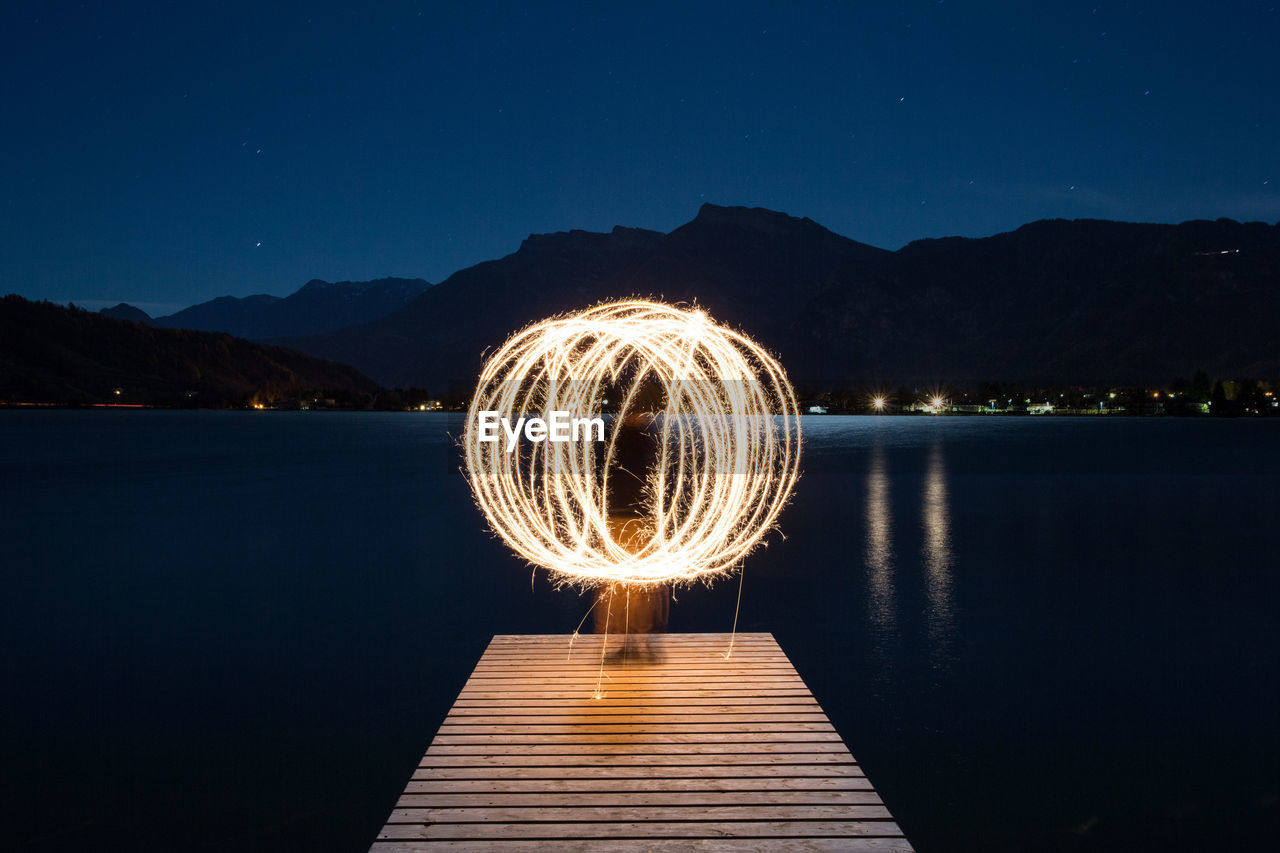 Person spinning illuminated wire wool while standing on pier against lake at night
