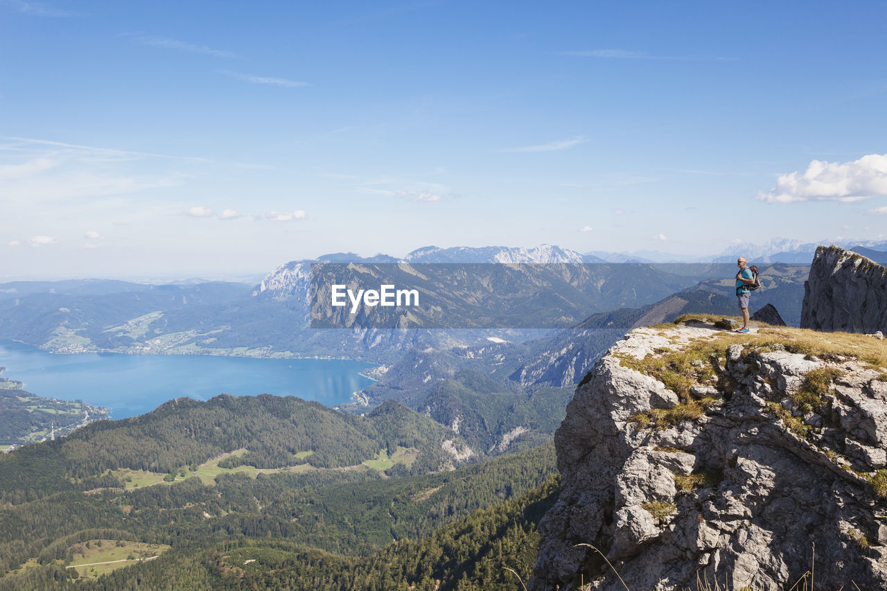 Full length of hiker standing on spinnerin peak while looking at attersee against blue sky