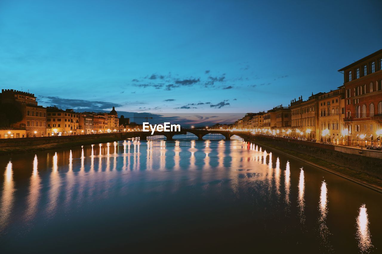 Bridge over river by illuminated buildings against sky at dusk