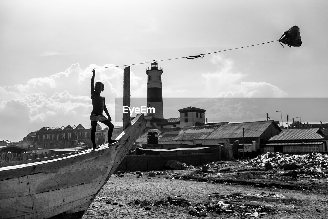 Shirtless boy holding kite while standing on boat against sky