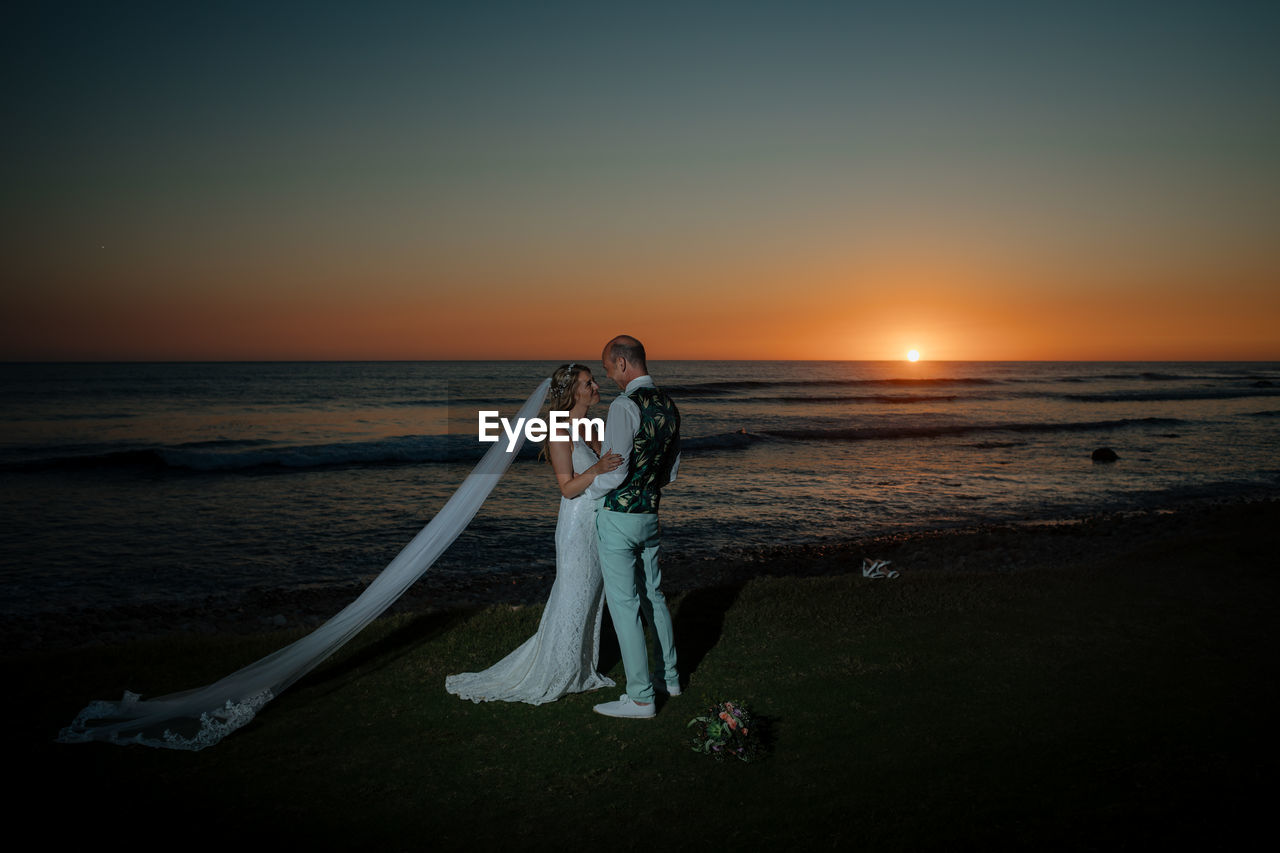 Wedding couple standing on beach against sky during sunset