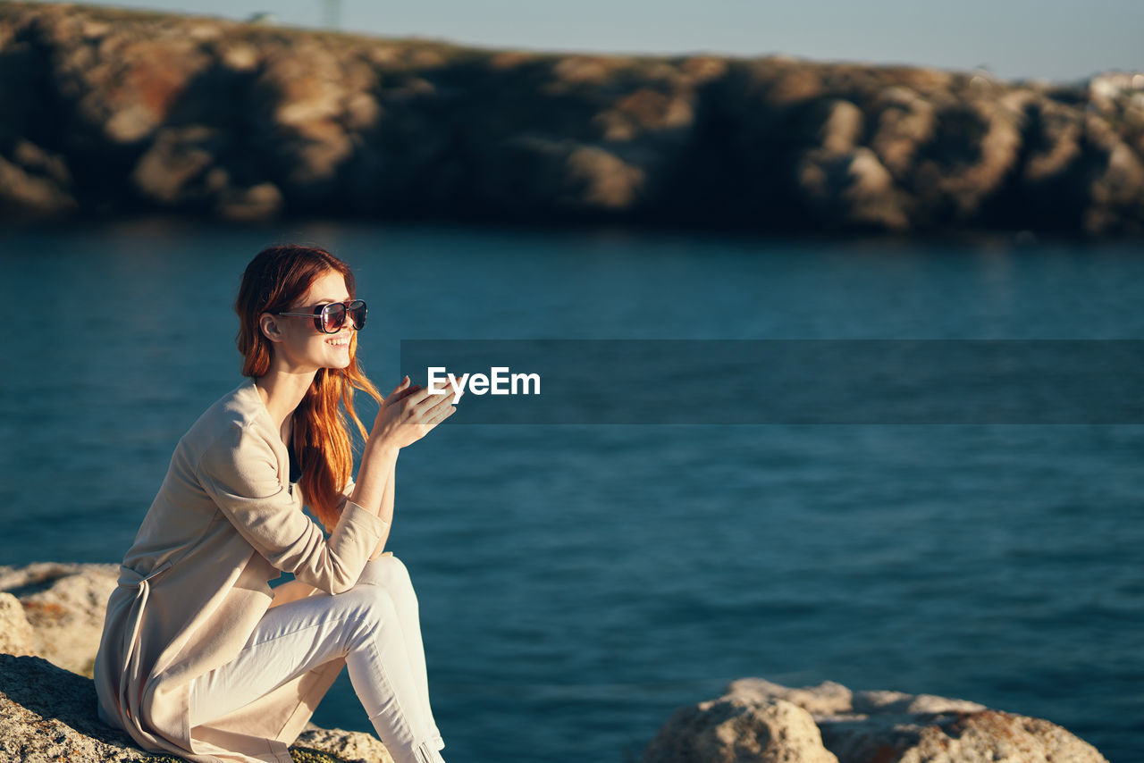 Young woman wearing sunglasses sitting on rock at sea shore