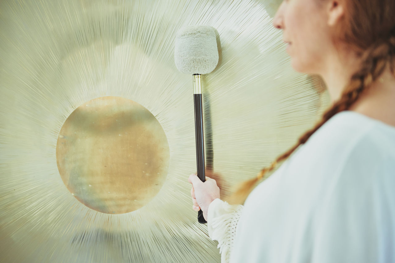 Side view of crop adult female in white clothes with mallet playing suspended gong during spiritual practice