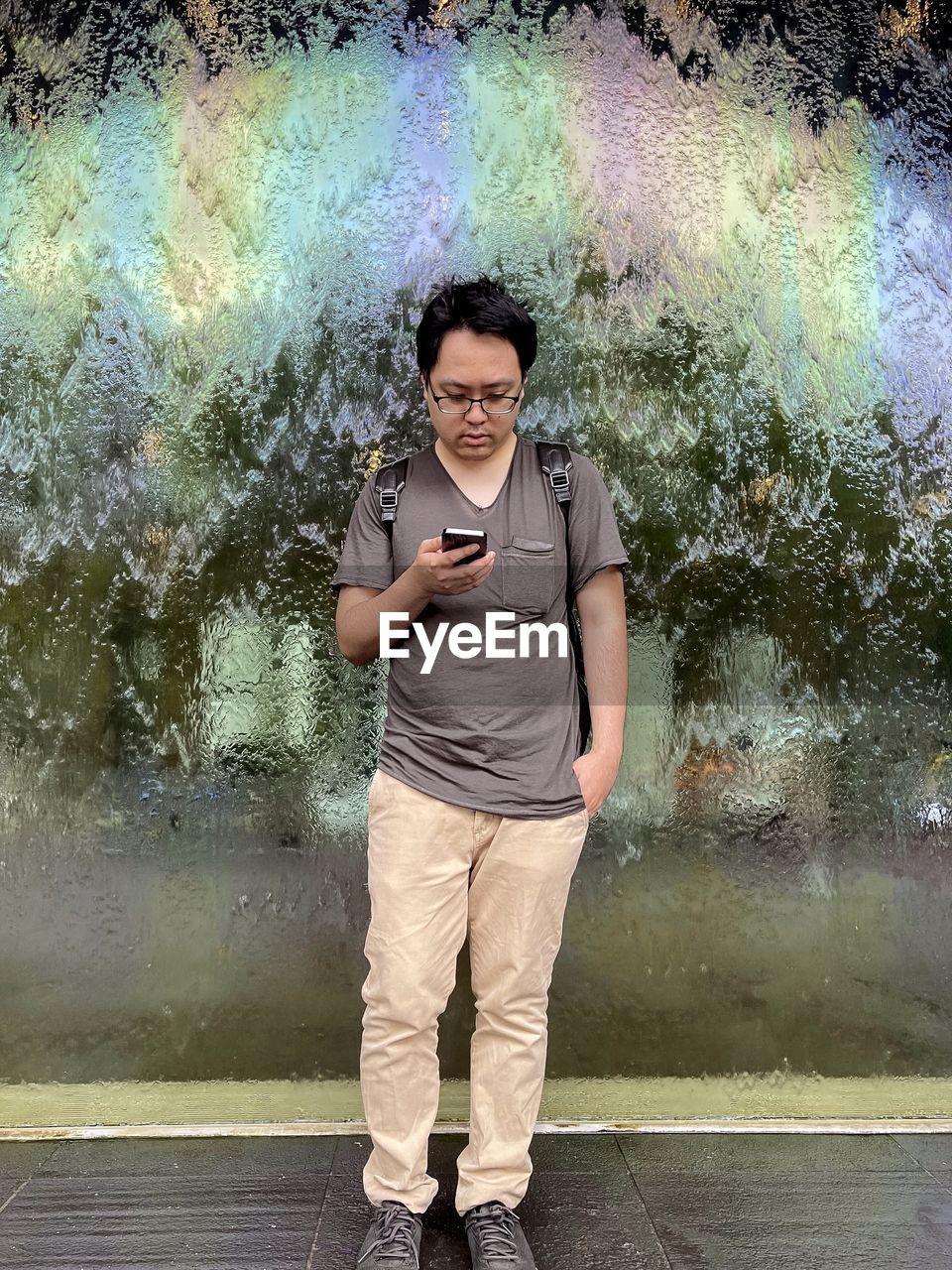 Portrait of young man using smartphone against rainbow patterned wet glass wall.