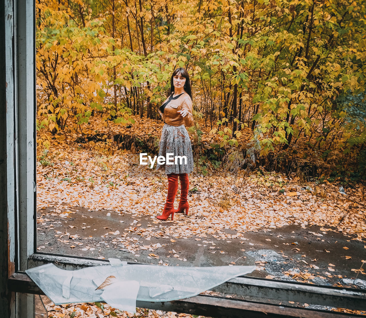 Full length portrait of woman against autumn trees on field seen through broken glass window