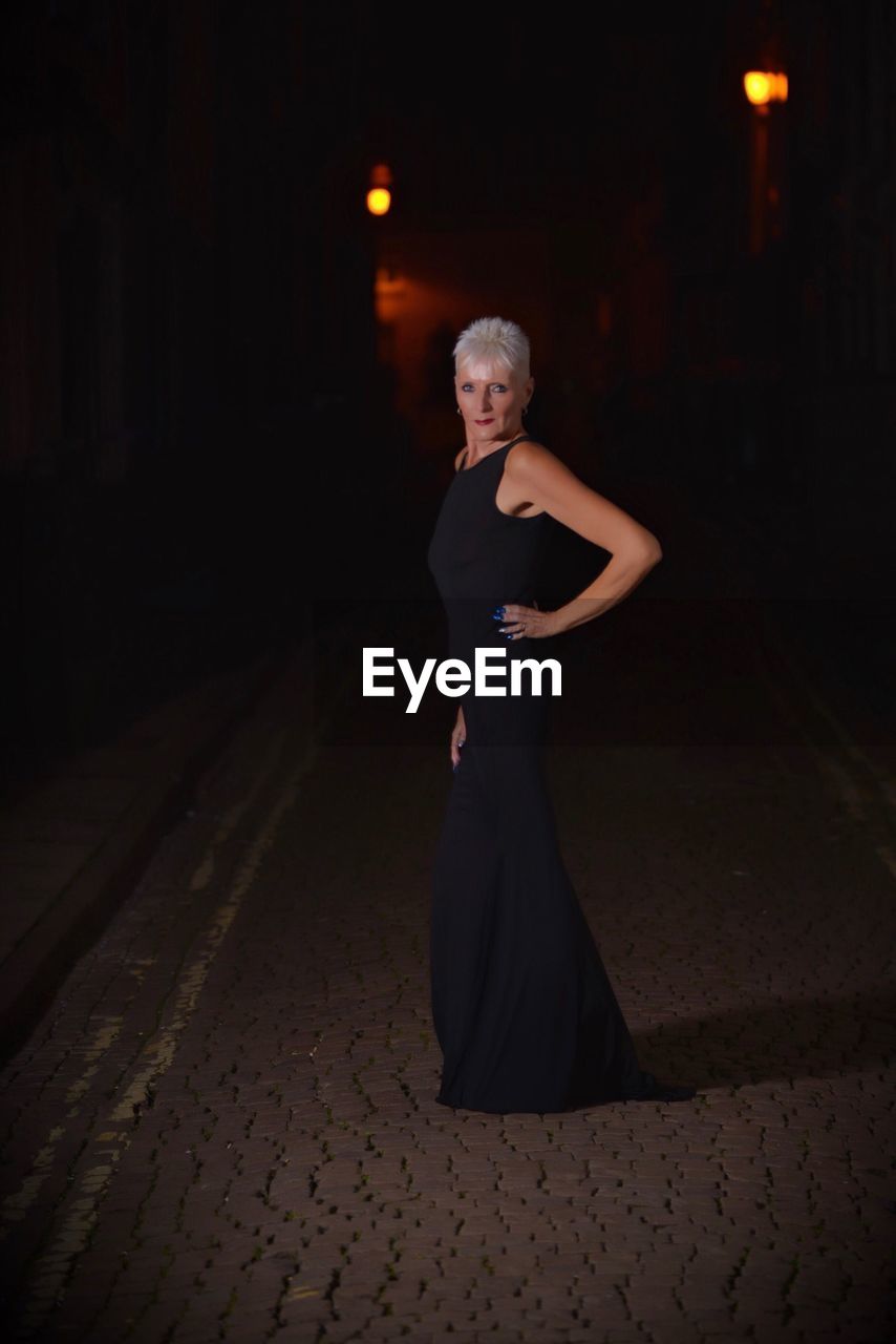 Portrait of confident mature woman wearing black dress standing on street at night