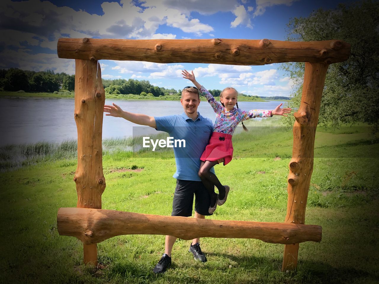 Father and daughter standing in front of wooden frame on field by lake