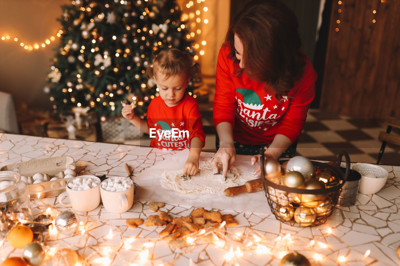 Parents and a child in red pajamas prepare christmas cookies in the decorated kitchen of the house