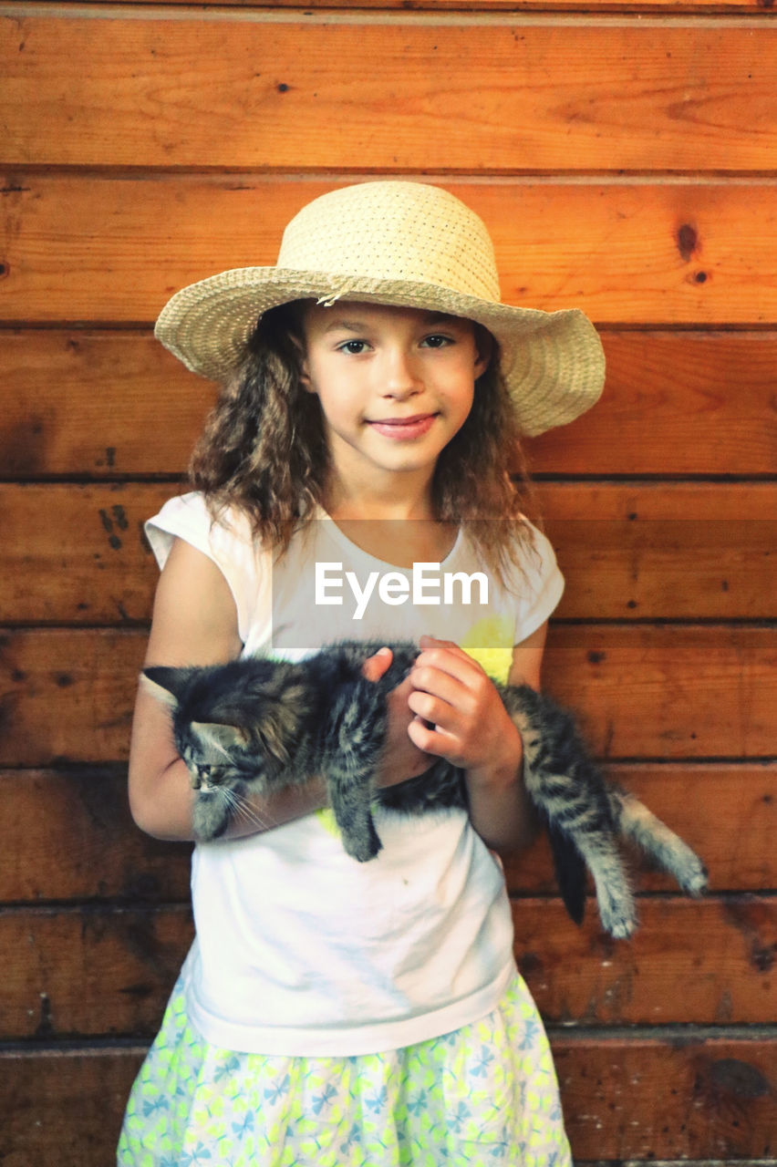Portrait of cute little girl wearing straw hat holding kitten standing against wooden planks in barn