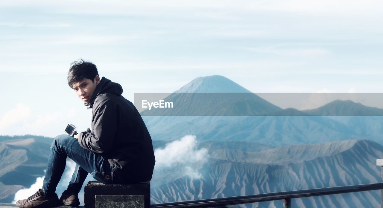 Side view portrait of man sitting on railing against volcano