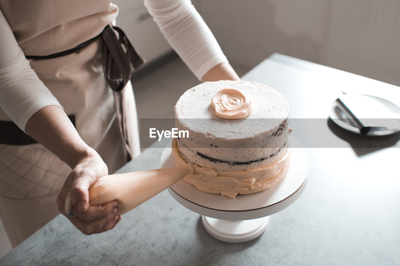 Woman holding pastry bag decorating cake with cream cheese standing in kitchen closeup.