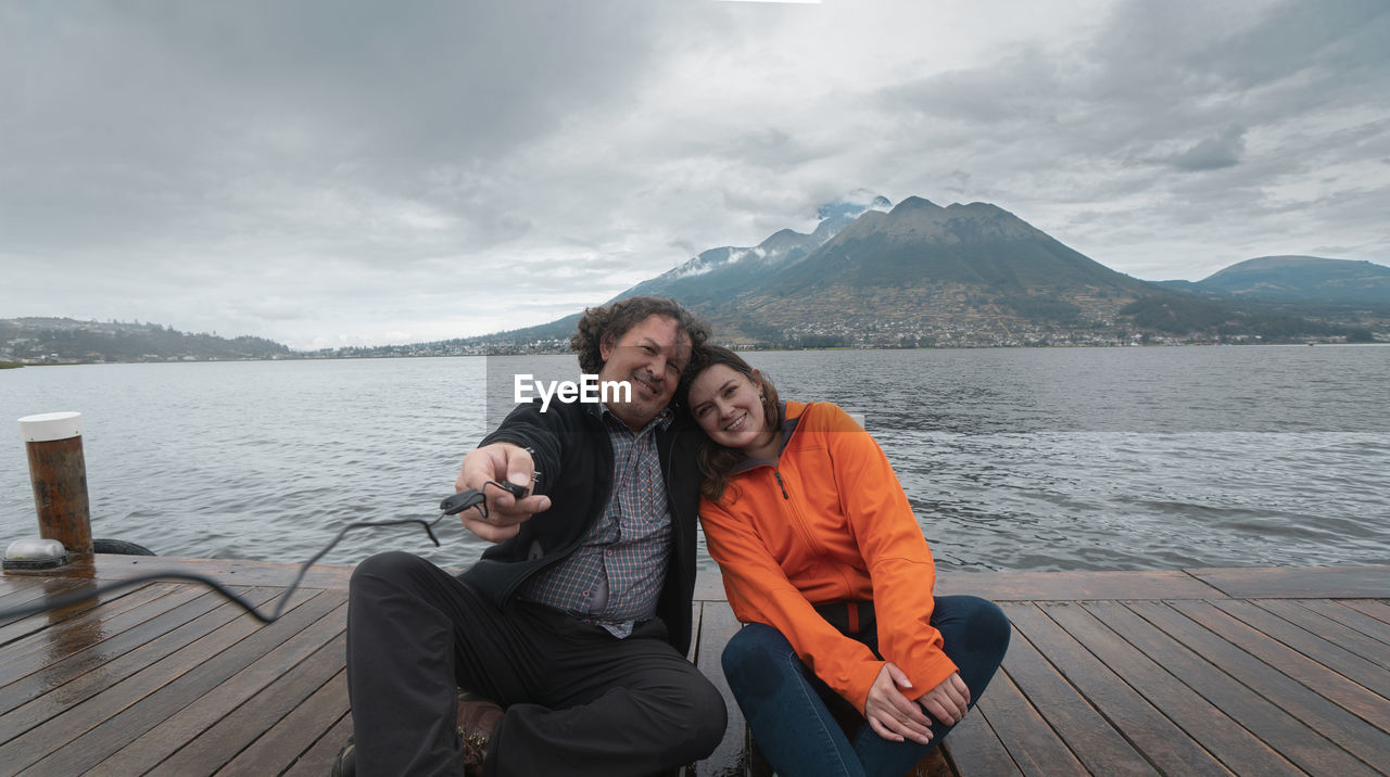 Young couple sitting on a wooden pier on lake san pablo taking a selfie with the imbabura volcano