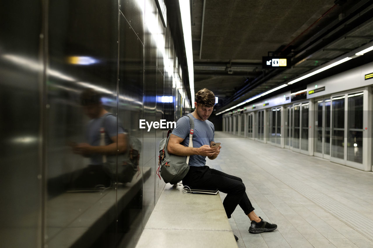 Young man using smart phone while sitting on bench against wall at illuminated subway