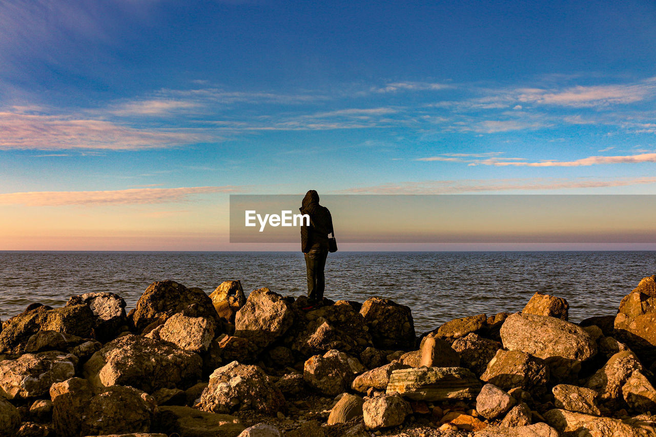 MAN STANDING ON ROCK IN SEA AGAINST SKY