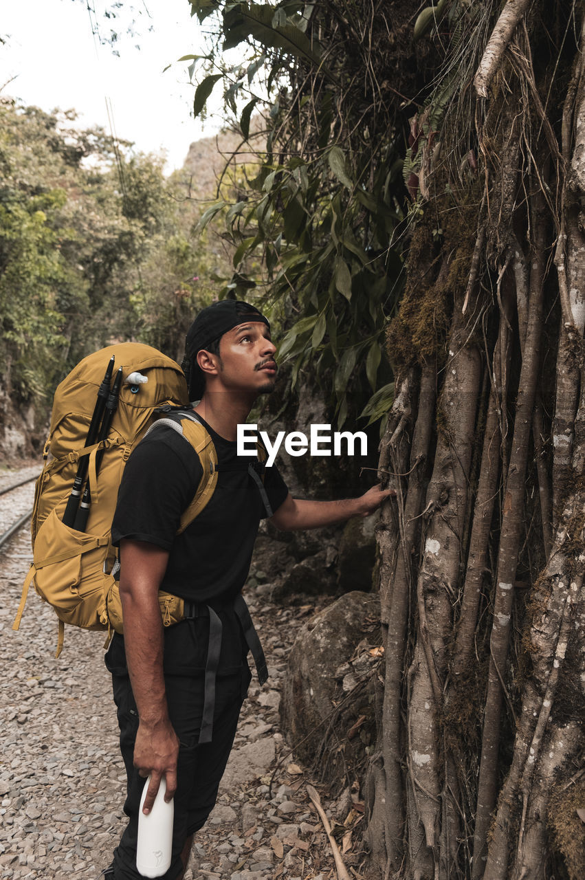 Side view of young man with backpack standing by tree in forest