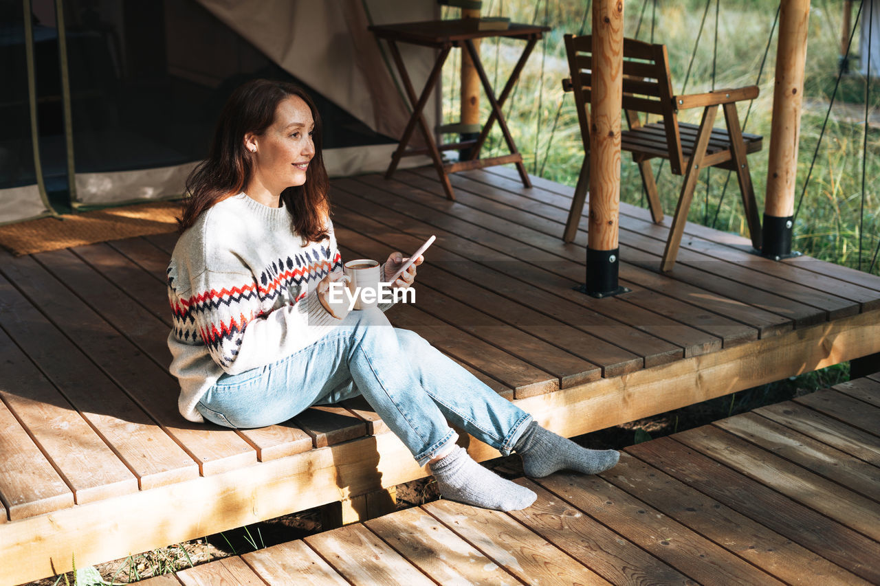 Brunette woman in nordic sweater using mobile phone and relaxing in glamping in nature