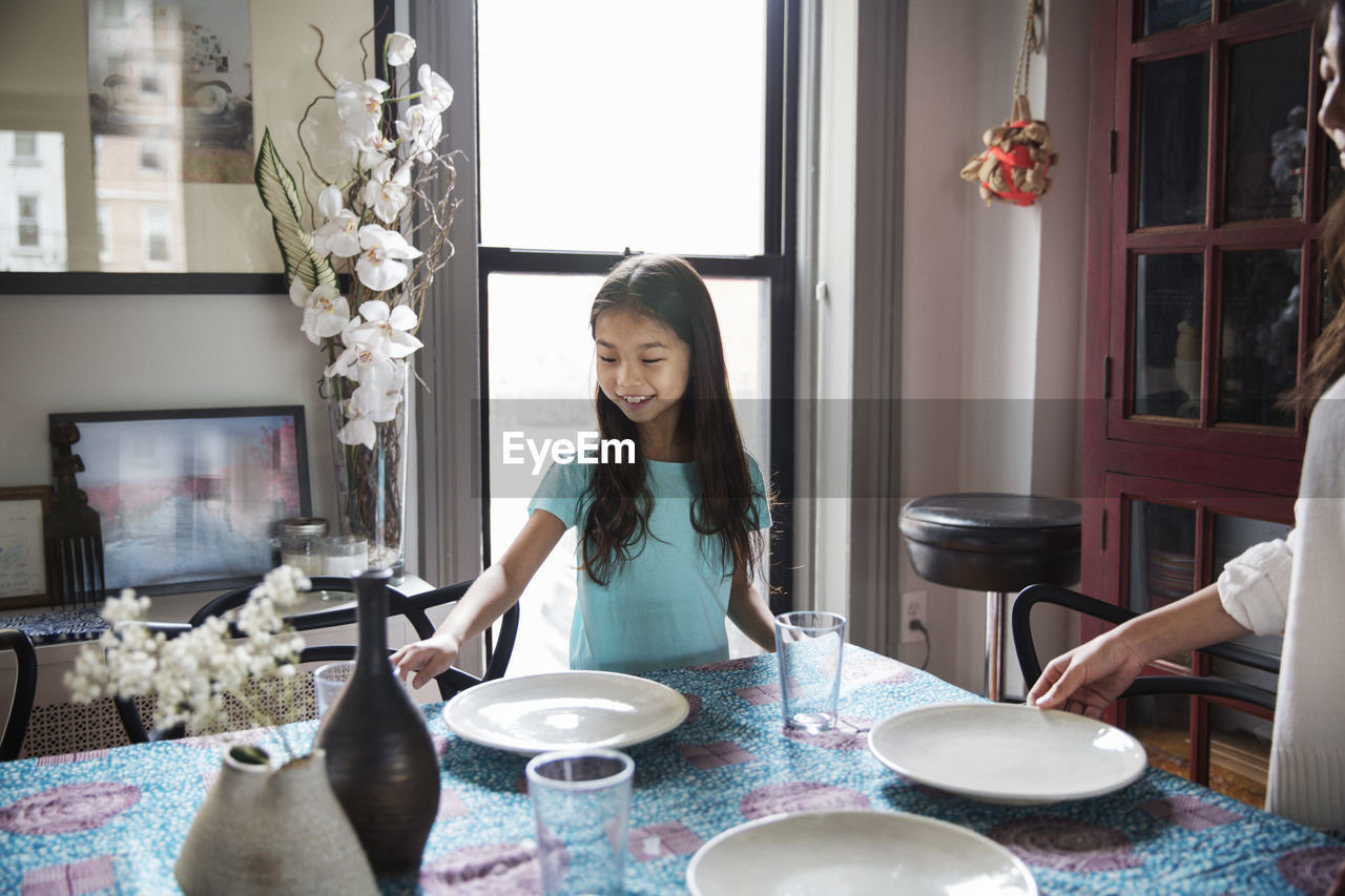 Happy sisters arranging plates on dining table at home