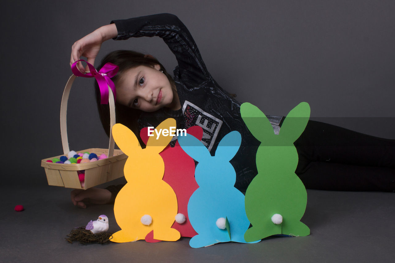 Portrait of smiling girl with eater bunny and pom pom in basket against gray background