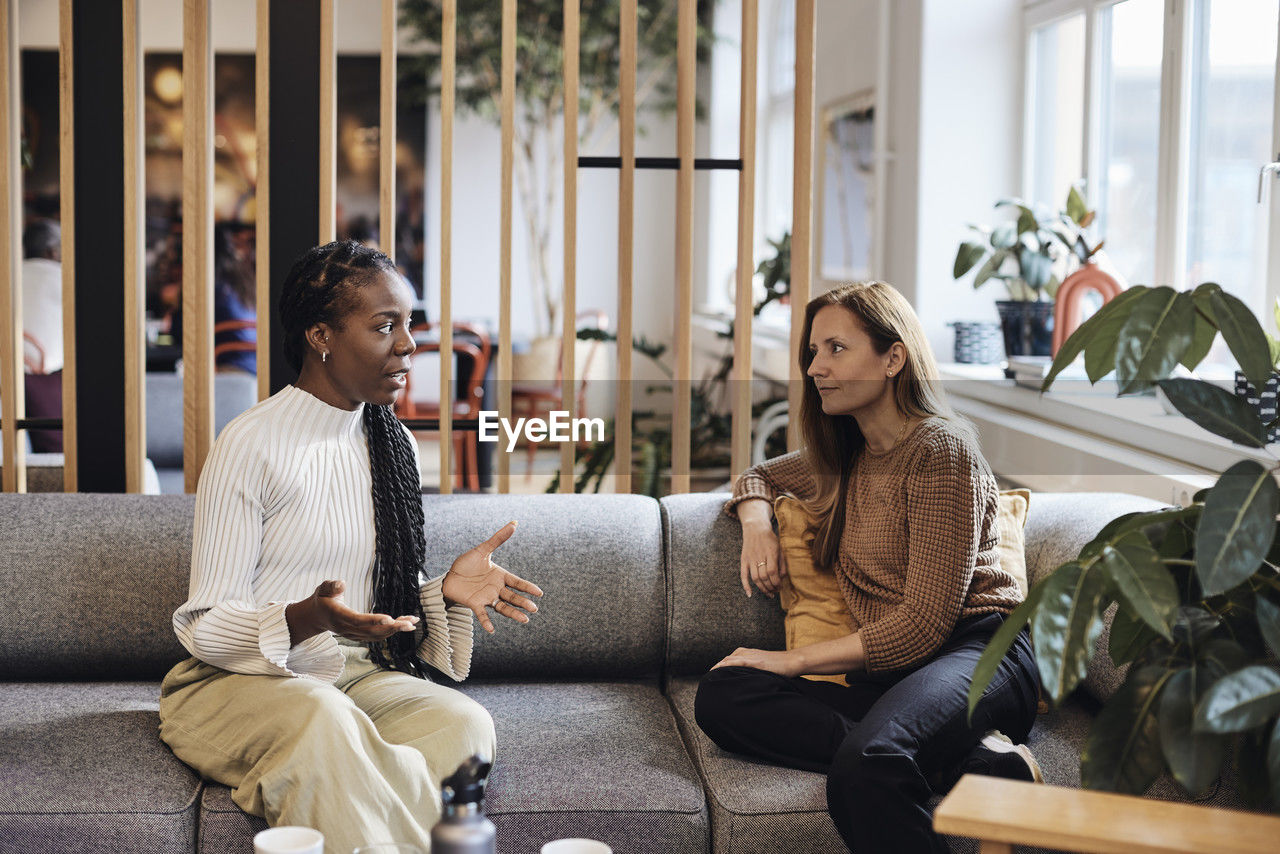 Multiracial female business colleagues discussing while sitting on sofa at coworking office