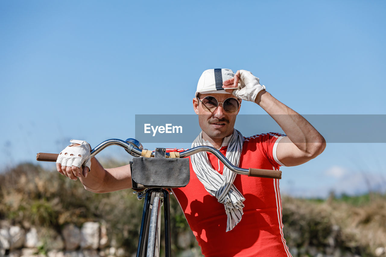 Stylish adult male with mustache in sunglasses and bright red shirt in composition with scarf and gloves touching striped cap while leaning on bike handlebar against blurred grassed hill and blue sky in nature