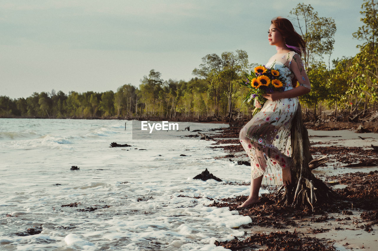 Full length of woman with flowers standing on sea shore against sky