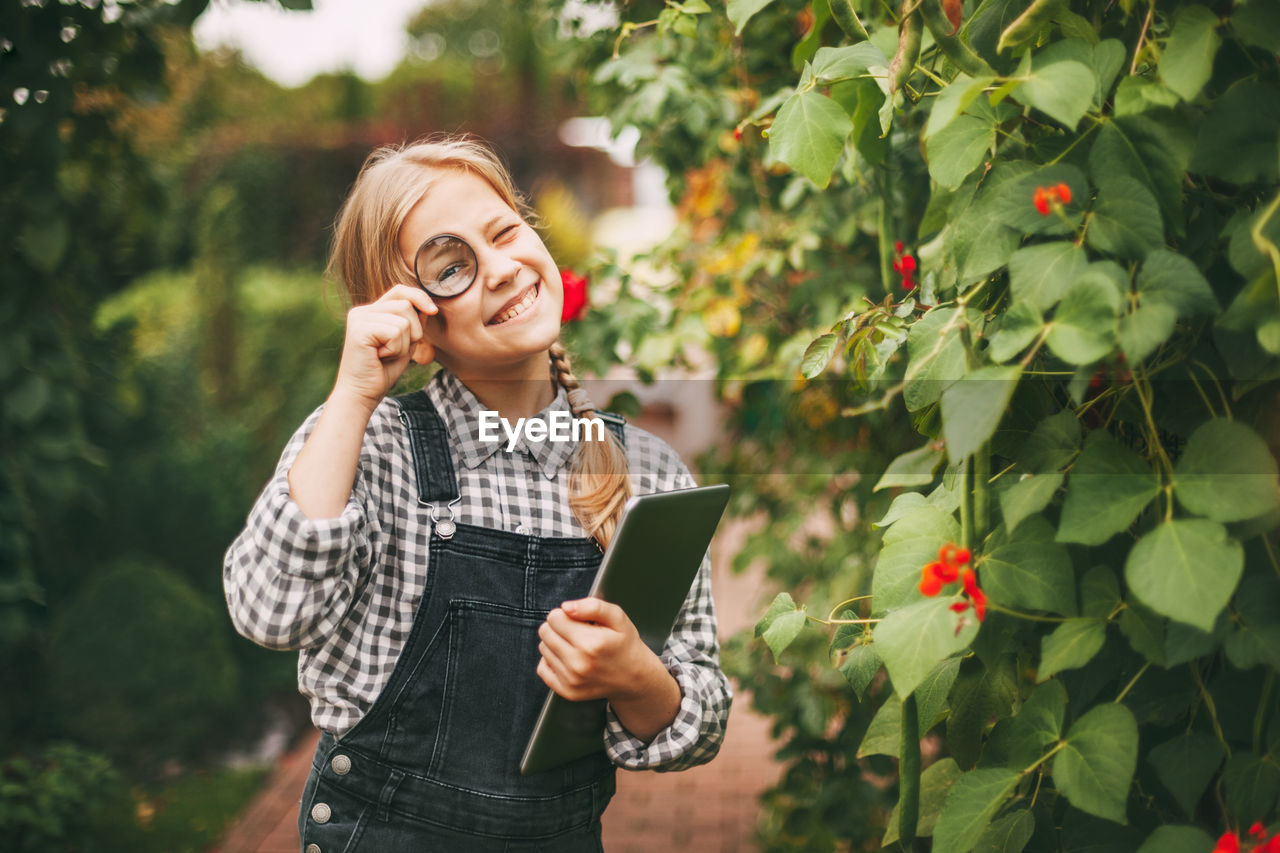 A beautiful little teenage girl with a tablet in her hands examines a sample of a plant through 