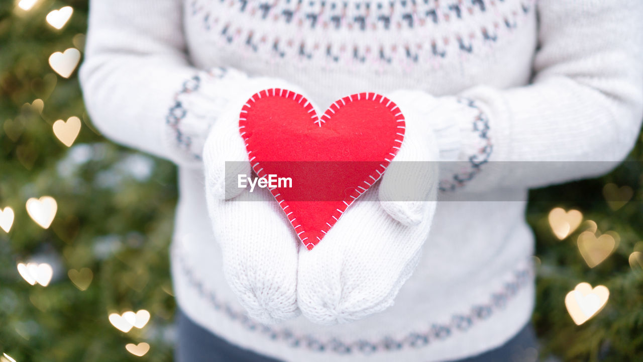 Red heart in the hands of a girl against the background of fir trees in gold bokeh. 