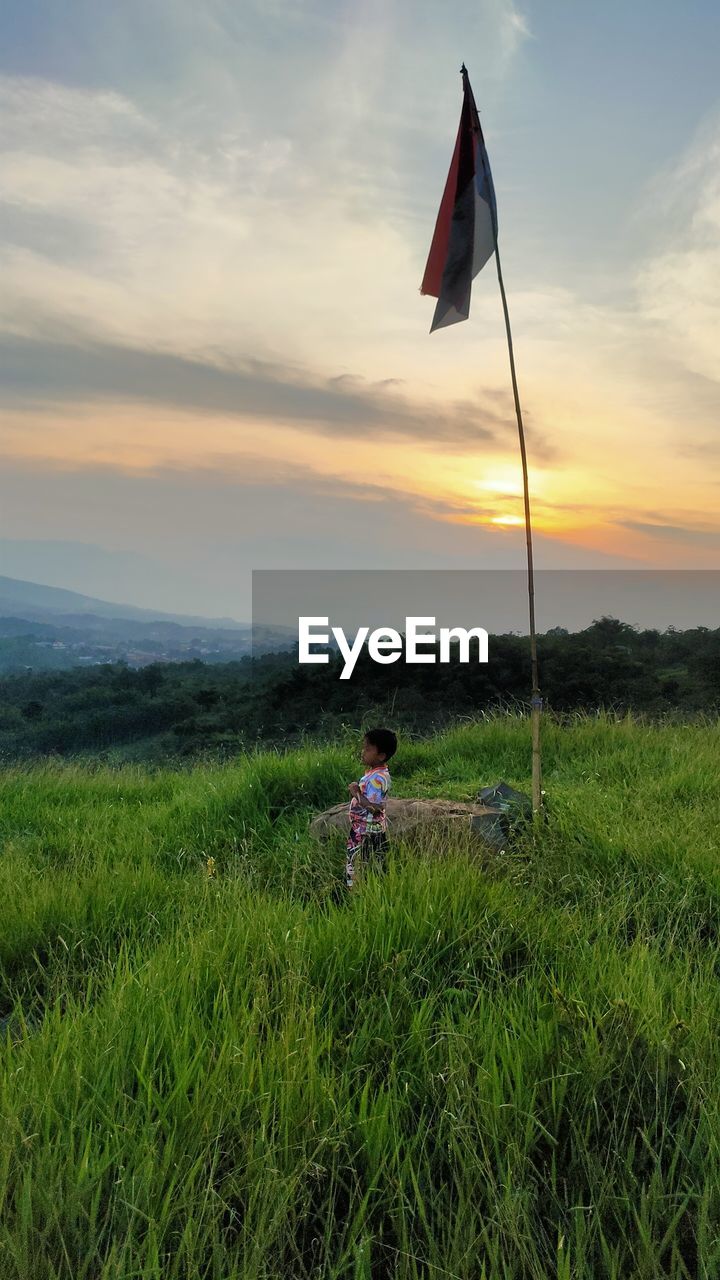 sky, grass, flag, plant, nature, cloud, field, environment, beauty in nature, grassland, land, one person, horizon, wind, sunset, leisure activity, meadow, landscape, green, prairie, rural area, scenics - nature, tranquility, natural environment, hill, tranquil scene, outdoors, lifestyles, activity, adult, patriotism, plain, day, men, child, childhood, rural scene, non-urban scene, women, growth