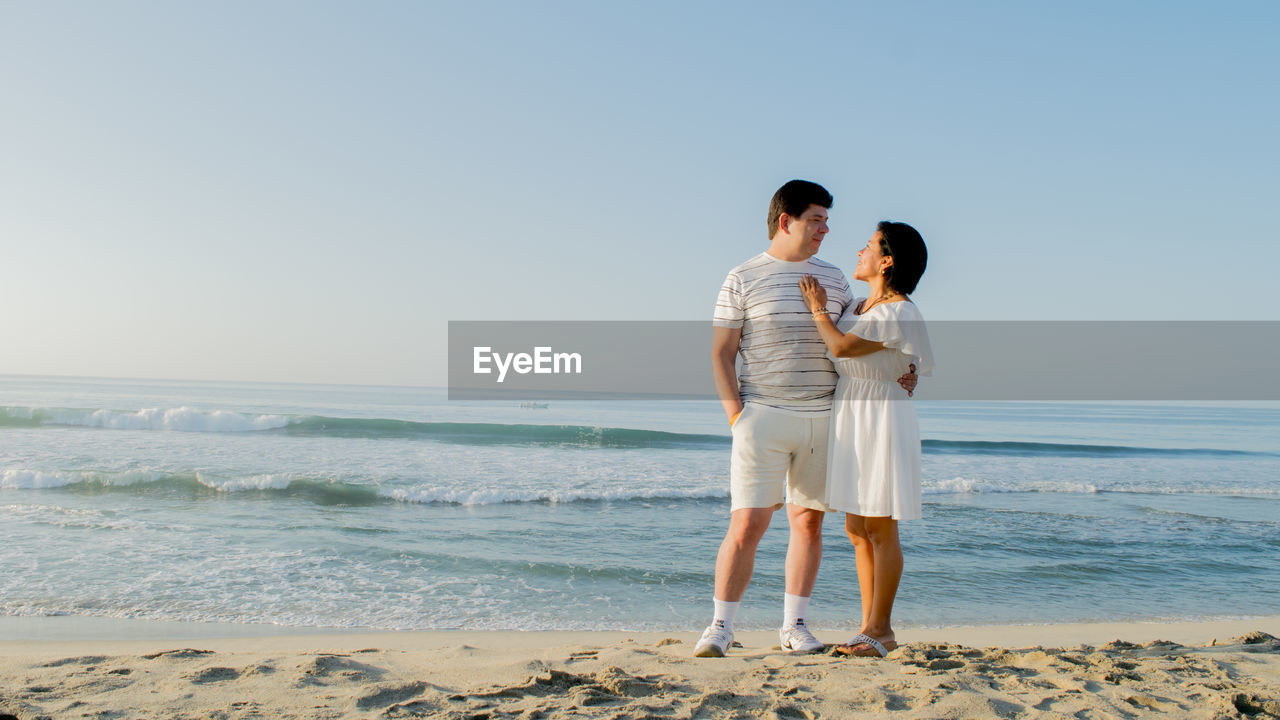 COUPLE STANDING AT BEACH AGAINST CLEAR SKY