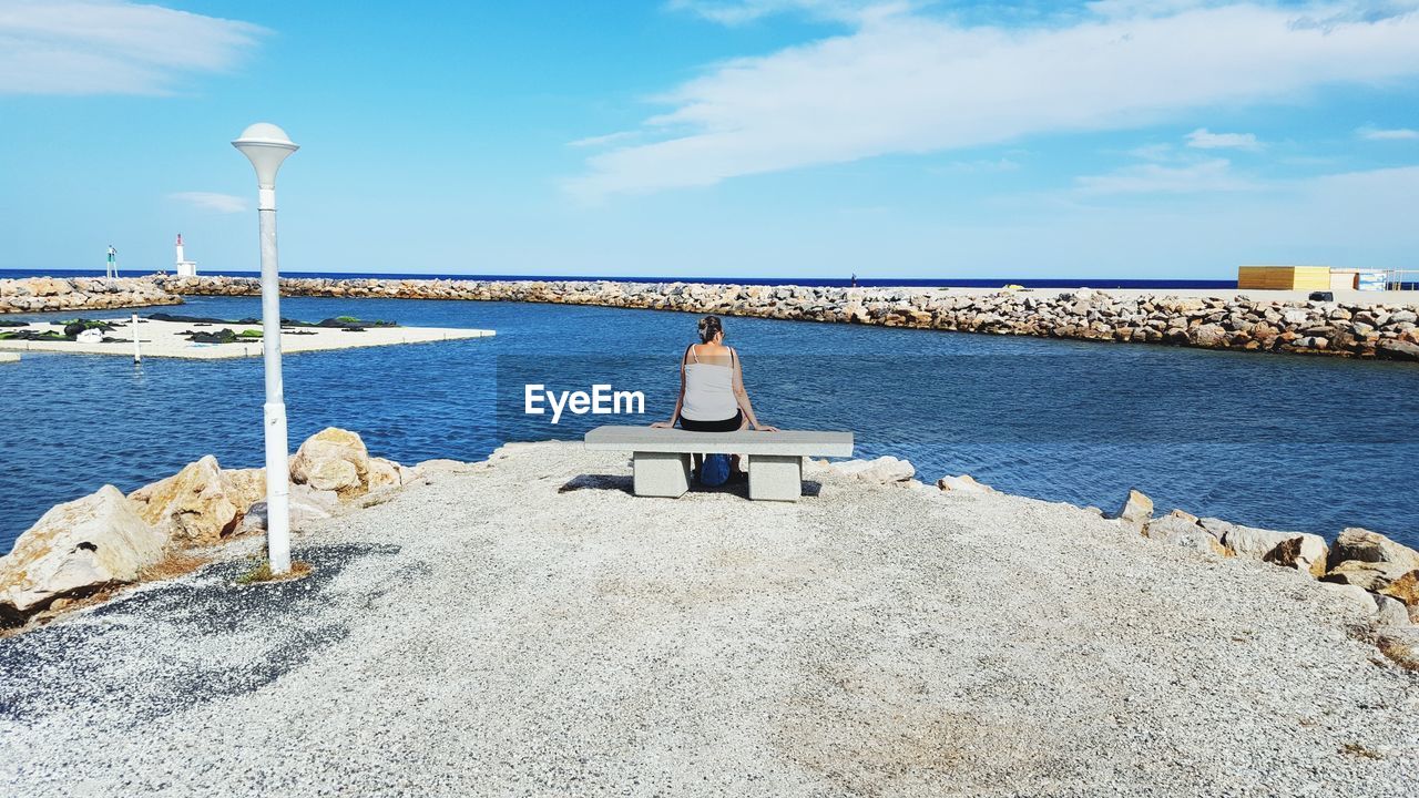 Rear view of mature woman sitting on bench at beach against sky during sunny day