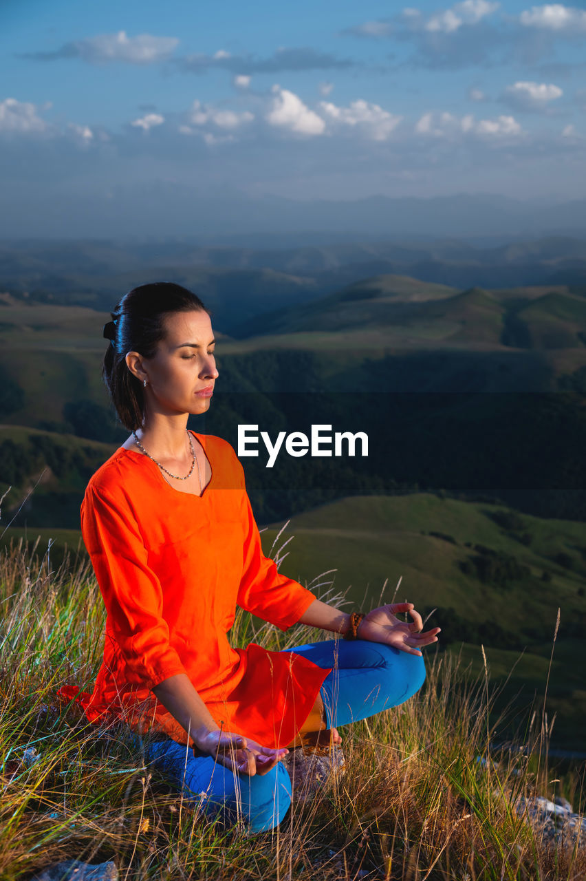Young yogi woman practicing meditation with closed eyes in lotus position at sunset
